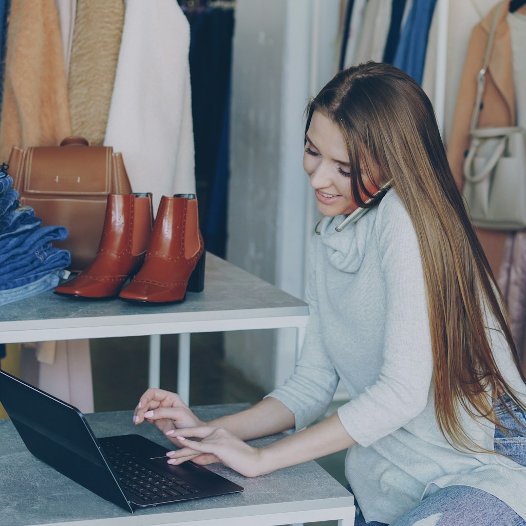 Owner of a small apparel brand taking a customer call at a laptop surrounded by stock